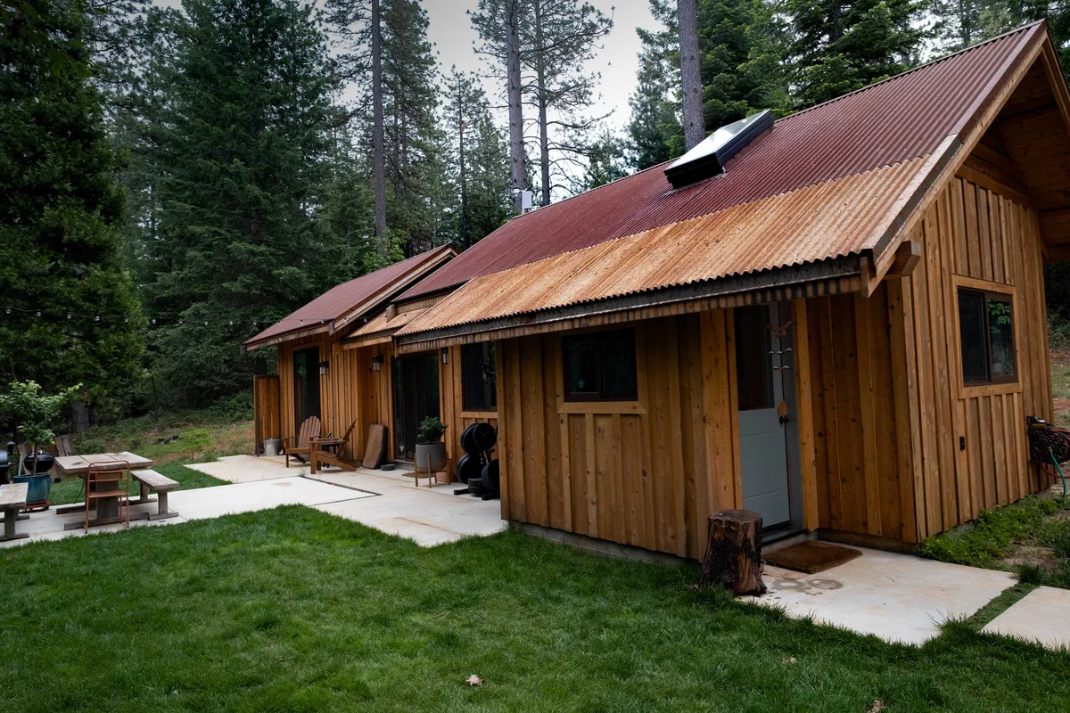 Two cedar-clad sheds joined into one cabin under weathered corrugated roofs, with a patio alongside
