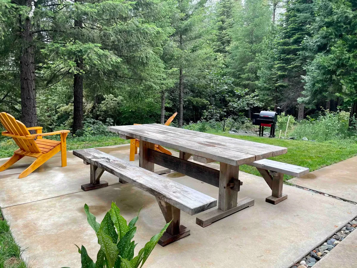 Long wooden picnic table and Adirondack chairs on a concrete patio at the forest edge